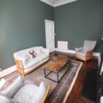 Cox & Co Estate agent in Edinburgh, Scotland. A cozy living room on Dundonald Street with dark green walls and white trim. It features a wooden coffee table, a beige sofa with cushions, and two matching armchairs. There is a patterned rug on the hardwood floor and a fireplace on the right side of the room.