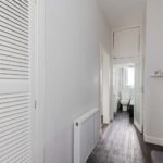 Cox & Co Estate agent in Edinburgh, Scotland. A white hallway with a closed louvered closet door on the left, dark wood flooring, a white radiator, and a bathroom at the end. The bathroom features a glass shower door, a pedestal sink, and a toilet. A small wall shelf is visible on the right – reminiscent of classic Edinburgh flats like those on Piersfield Terrace in EH8.