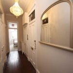 Cox & Co Estate agent in Edinburgh, Scotland. A narrow hallway on Dundonald Street with dark wooden flooring and light-colored walls features white doors on the right side. A large, gold-framed mirror is mounted on the right wall, and an ornate chandelier hangs from the ceiling. Natural light filters in from the far end.