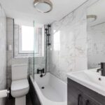 Cox & Co Estate agent in Edinburgh, Scotland. A modern bathroom in Piersfield Terrace, Edinburgh, featuring a white toilet, a bathtub with a glass divider, a black shower fixture, and a sink with a dark wood cabinet below a large mirror. The walls are covered with white marble tiles, and there is a round light fixture on the ceiling.
