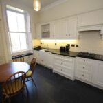 Cox & Co Estate agent in Edinburgh, Scotland. A kitchen on Dundonald Street with white cabinetry, a black countertop, and a white tile backsplash. It features a gas stove, dishwasher, and various small appliances. A round wooden table with four chairs sits near a large window, and the floor is tiled in dark gray.