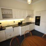 Cox & Co Estate agent in Edinburgh, Scotland. A spacious kitchen on Dundonald Street features cream-colored cabinets, black countertops, and a farmhouse sink. The kitchen is equipped with a gas stovetop, built-in oven, and microwave. A round wooden table with four chairs is placed in the center. The lighting is warm and inviting.