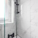 Cox & Co Estate agent in Edinburgh, Scotland. A modern bathroom with white marble tile walls featuring a black showerhead attached to a black vertical bar and a black faucet mounted on the side of a white bathtub. A glass partition is visible on the left, next to a window allowing natural light to fill this elegant space in Edinburgh's EH8 district.