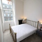 Cox & Co Estate agent in Edinburgh, Scotland. A minimalist bedroom on Dundonald Street with a large window letting in natural light. The room features a double bed with a white quilted mattress and a brass headboard, flanked by two bedside tables with matching lamps. The walls are painted white, and there is an empty fireplace.