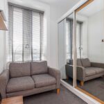 Cox & Co Estate agent in Edinburgh, Scotland. A small room in EH8, featuring a grey sofa set against the wall under a large window with blinds. A shelf is mounted on the adjacent wall, and a mirrored sliding door closet on the right reflects the room and sofa. A small wooden table sits next to the sofa, completing this cozy Edinburgh space on Piersfield Terrace.