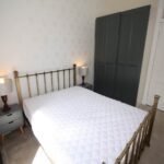 Cox & Co Estate agent in Edinburgh, Scotland. A cozy bedroom on Dundonald Street features a double bed with a white quilt, flanked by bedside tables and lamps. A dark green wardrobe stands opposite a white door, complementing the light-colored carpet and wallpaper.
