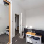 Cox & Co Estate agent in Edinburgh, Scotland. A small, well-lit bedroom on Piersfield Terrace, Edinburgh, with a gray bed against a white wall. A wooden side table with drawers and electronic devices is beside the bed. An open door leads to an adjacent room. A bookshelf is mounted on the wall, and a mirror reflects part of the other room.