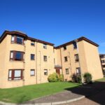 Cox & Co Estate agent in Edinburgh, Scotland. A three-story, beige brick apartment building with brown-framed windows, set against a clear blue sky in the West Powburn area. Small shrubs and green grass surround the entrance, with a paved walkway leading to the door.