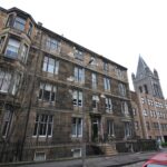 Cox & Co Estate agent in Edinburgh, Scotland. A street view of a row of multi-story buildings with brick facades. The left building features bay windows, with leafless trees in front. A lamp post is in the center, and on the right is a taller, more modern Place Leslie building with a pointed tower. A red car is partially visible at the bottom right.