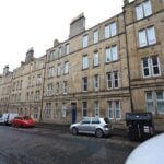 Cox & Co Estate agent in Edinburgh, Scotland. A street view of Yeaman Place in an urban area featuring a row of beige stone apartment buildings with multiple rectangular windows. Several parked cars and a dumpster line the street, which appears to be wet, indicating recent rain. The sky is overcast.
