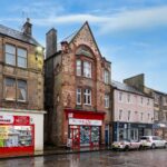 Cox & Co Estate agent in Edinburgh, Scotland. A rainy day on Midlothian’s High Street reveals a street lined with buildings. The central structure boasts a stone facade, arched windows, and a "Coral" sign. To its left is the Dalkeith Home & Hardware store, while a brick building stands to the right amid parked cars.
