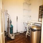 Cox & Co Estate agent in Edinburgh, Scotland. A small, narrow room on Yeaman Place with wooden flooring. There is a vacuum cleaner and a mop stored on one side, and a desk with two folding chairs on the other. Above the desk, shelves hold various items. A trash can is placed next to the desk.