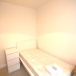 Cox & Co Estate agent in Edinburgh, Scotland. A small, sparsely furnished bedroom with a single bed covered in a white mattress and pillow. Next to the bed is a white three-drawer dresser, and a wooden nightstand is positioned on the other side. The walls are painted white, and there is a closed wooden door to place your belongings securely.