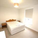 Cox & Co Estate agent in Edinburgh, Scotland. A minimalist bedroom at Ferry Gait Place features a double bed with a wooden headboard and a white mattress. Two wooden nightstands frame the bed. The room has a single window with a white roller shade, beige carpet, and a ceiling light with a beige lampshade.