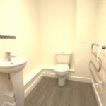 Cox & Co Estate agent in Edinburgh, Scotland. A minimalistic bathroom in Ferry Gait Place features a white sink with a silver faucet on the left and a white toilet next to it. The floor is covered with grey tiles, and a towel rack is mounted on the right wall. The walls are painted white, creating a clean and bright atmosphere.
