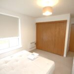 Cox & Co Estate agent in Edinburgh, Scotland. A minimalist bedroom with neutral tones features a single bed with a white mattress and folded towel, a small white dresser, a large wooden closet with sliding doors, a window with a roller blind, and a wooden door leading to another room. A ceiling light is on, casting a gentle glow over this tranquil place.