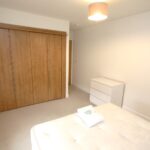 Cox & Co Estate agent in Edinburgh, Scotland. A minimalistic bedroom with a double bed in the foreground, topped with white linen and a folded towel. A wooden wardrobe with sliding doors is placed along one wall, and a white dresser stands adjacent to the bed. This serene place is illuminated by a ceiling light fixture.
