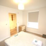 Cox & Co Estate agent in Edinburgh, Scotland. A minimalist bedroom with a single bed, covered with a mattress, pillow, and folded towel at the foot. The room features white walls, a tall window with a blind, a wooden dresser, and a small black table. An adjacent bathroom is visible through an open door—an ideal place to unwind after disembarking the ferry.