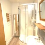 Cox & Co Estate agent in Edinburgh, Scotland. A small bathroom at Ferry Gait Place features a glass-enclosed corner shower with a chrome showerhead, a white sink with a chrome faucet, and a mirror with a wooden frame above the sink. A wooden door, wall-mounted towel rack, and full-length mirror complete this tidy space.