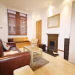 Cox & Co Estate agent in Edinburgh, Scotland. A cozy living room on Yeaman Place features a leather sofa, a wooden coffee table, a fireplace with a framed picture above, and a window with wooden blinds. The room has wooden floors, a beige rug, modern lighting, and a built-in storage bench under the window.