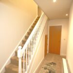 Cox & Co Estate agent in Edinburgh, Scotland. A well-lit hallway with a carpeted staircase on the left featuring white balusters and railings. The hallway, much like a ferry place of transit, has beige walls, a wooden door at the end on the right, and a small patterned rug on the floor. Recessed ceiling lights illuminate the space.