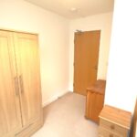 Cox & Co Estate agent in Edinburgh, Scotland. A small bedroom with plain white walls features a wooden wardrobe, a bed with a wooden frame, and a small wooden nightstand. There's a wooden door in the background leading out of the room, next to a small wooden chest. The floor is carpeted in light beige, giving the place a cozy feel.