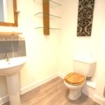Cox & Co Estate agent in Edinburgh, Scotland. A small bathroom with light-colored walls features a pedestal sink with a mirror above it on the left, a toilet with a wooden seat on the right, and a towel rack to the side. Three glass shelves are mounted on the wall above the toilet, and a decorative tree art piece graces this serene place.