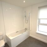 Cox & Co Estate agent in Edinburgh, Scotland. A modern bathroom at Ferry Gait Place features a white bathtub and shower, a tiled backsplash, and a toilet in the left corner. The window on the right has a roller shade partially down, letting light in. The floor appears to be covered with gray vinyl or laminate.