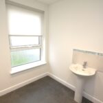 Cox & Co Estate agent in Edinburgh, Scotland. A bright, minimally furnished bathroom with a single frosted window and a white roller blind. The room features a pedestal sink with a tiled backsplash and a tiled area to the left, suggesting the presence of a bathtub or shower. The floor is covered with dark vinyl, giving the place an elegant gait.