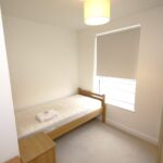 Cox & Co Estate agent in Edinburgh, Scotland. A small, brightly lit bedroom at Ferry Gait Place features white walls with a single bed against the left wall. The bed has a white mattress and a folded towel on top. Next to it is a small wooden bedside table, while a tall wooden wardrobe stands to the right. A window with a closed blind is on the far wall, illuminated by a ceiling light.