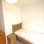 Cox & Co Estate agent in Edinburgh, Scotland. A simple bedroom with a single bed against a white wall. The bed has a wooden frame, and the mattress is bare except for a folded towel and a small item on top. A wooden chest of drawers and a small bedside table are placed next to the bed. The room is brightly lit, creating an inviting place to relax.