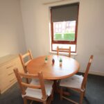 Cox & Co Estate agent in Edinburgh, Scotland. A small dining area in West Powburn features a round wooden table and four wooden chairs, each with a white cushion. The table is adorned with three small potted plants for decoration. A light-colored dresser stands against the left wall, and a window offers an outside view of the surrounding buildings.