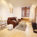 Cox & Co Estate agent in Edinburgh, Scotland. A cozy, well-lit living room on Yeaman Place featuring a brown leather sofa, a wooden coffee table on a beige rug, a wicker chair, and a wooden side table with a lamp. The room is adorned with white walls, wooden blinds on the window, and a fireplace with a mirror above it on the right.