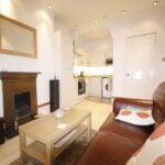 Cox & Co Estate agent in Edinburgh, Scotland. A cozy living room on Yeaman Place features a brown leather sofa adorned with white and orange pillows, facing a wooden coffee table. A fireplace with a mirror above it is on the left wall. An open-plan kitchen with white cabinets and modern appliances is visible in the background.
