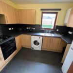 Cox & Co Estate agent in Edinburgh, Scotland. A modern kitchen in West Powburn featuring light wood cabinets, a black countertop, and black tiled flooring. Appliances include a gas stove, oven, washing machine, and refrigerator. A window with a view of neighboring houses is centered above the sink area.