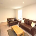 Cox & Co Estate agent in Edinburgh, Scotland. A living room at Ferry Gait with two brown leather sofas, one smaller and one larger, each adorned with white pillows. In front of the sofas, there is a wooden coffee table on a gray rug. The room is well-lit by a window with white blinds and features beige walls and carpet.