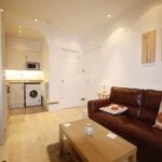 Cox & Co Estate agent in Edinburgh, Scotland. A cozy living room on Yeaman Place features a brown leather sofa and a light wood coffee table. The adjacent compact kitchen has white cabinets, a washing machine, microwave, and warm lighting. The walls are adorned with two minimalist art pieces, and the floor is light wood laminate.