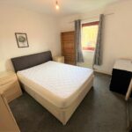 Cox & Co Estate agent in Edinburgh, Scotland. A tidy bedroom in West Powburn features a neatly made double bed with a white quilt and dark headboard. Light wooden nightstands flank the sides, accompanied by an empty wooden bookcase, black dresser, desk lamp, and window with gray curtains. A framed picture adorns the wall.