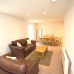 Cox & Co Estate agent in Edinburgh, Scotland. A spacious living room at Ferry Gait with beige walls, featuring two brown leather sofas, a wooden coffee table, and a matching area rug. In the background, there is a dining area with a wooden table and six chairs. The room is well-lit with recessed ceiling lights and natural light from windows on the left.