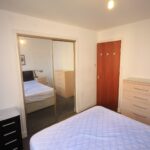 Cox & Co Estate agent in Edinburgh, Scotland. A simple bedroom in West Powburn with a bed, a wooden dresser, and a door on the right. A large mirror on the left side of the room reflects part of the bed and furniture. The walls are white, and there's a corkboard on one wall. The lighting comes from a single ceiling lamp.