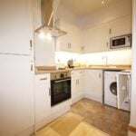 Cox & Co Estate agent in Edinburgh, Scotland. A modern, compact kitchen at Yeaman Place features white cabinets, light brown countertops, and tiled flooring. It includes an oven, stovetop, extractor fan, microwave, sink, and a washing machine. Recessed lighting illuminates the workspace, complemented by a small mat on the floor.
