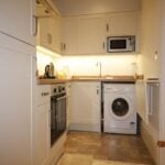 Cox & Co Estate agent in Edinburgh, Scotland. A compact, modern kitchen at Yeaman Place features cream-colored cabinets, an oven, a cooktop, a washing machine, a microwave, and a sink. The countertops are clutter-free, and the floor is tiled with a beige pattern. Warm under-cabinet lighting beautifully illuminates the space.