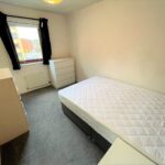 Cox & Co Estate agent in Edinburgh, Scotland. A cozy West Powburn bedroom featuring a bed with a white quilted mattress, a black fabric headboard, two white dressers with drawers, and a window dressed in black curtains. The room's white walls and gray carpeting are bathed in natural light.