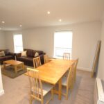 Cox & Co Estate agent in Edinburgh, Scotland. A spacious, well-lit living and dining area with white walls. The dining area features a wooden dining table with six chairs. The living space is the perfect place to relax with two brown sofas, a coffee table, and carpeting. Two large windows ferry natural light into the room.