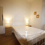 Cox & Co Estate agent in Edinburgh, Scotland. A serene bedroom on Yeaman Place features a white metal-framed double bed covered in a plastic sheet, flanked by two wooden nightstands each with a lit lamp. A tall mirror is mounted on the wall to the left, and two small framed pictures are hung on the right wall adjacent to a large window.