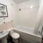 Cox & Co Estate agent in Edinburgh, Scotland. A compact, modern bathroom in West Powburn features a white bathtub and shower curtain on the right, a white toilet and sink on the left, and a framed botanical print above the sink. The white walls are complemented by a sleek glass shelf above the sink.