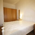 Cox & Co Estate agent in Edinburgh, Scotland. A cozy bedroom at Yeaman Place features a double bed with an iron frame and a mattress without bedding. On either side of the bed are wooden bedside tables with lit lamps. A large wardrobe with sliding wooden doors is built into the wall, and a small framed picture hangs next to a radiator.