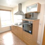 Cox & Co Estate agent in Edinburgh, Scotland. A modern kitchen with light wood cabinetry and beige tiled floor. It features a built-in oven, a microwave on top, a stovetop with an overhead stainless steel range hood, and a washing machine to the left of the stove. There is a window with a brown blind, making it the perfect place for culinary creativity.
