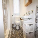 Cox & Co Estate agent in Edinburgh, Scotland. A small, white-tiled bathroom on Yeaman Place features a pedestal sink and a toilet. A wicker storage unit is placed beneath the sink, while a heated towel rail graces the right side. The floor is adorned with small, mosaic-patterned tiles and a frosted window sits above the toilet.