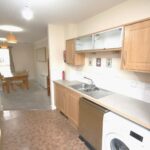 Cox & Co Estate agent in Edinburgh, Scotland. A modern, narrow kitchen at Ferry Gait Place features light wooden cabinets and a light brown tiled floor. Appliances include a dishwasher, washing machine, and microwave. The kitchen opens into a dining area with a wooden table and chairs, illuminated by natural light through the window.